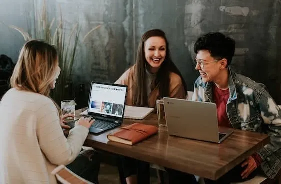 Tres personas sonriendo mientras trabajan con portátiles en una mesa de madera